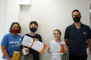 Aluno, aluna, professor e professora estão posicionados a frente de uma parede branca. Os alunos estão no meio dos professores e seguram os certificados do Concurso de Leitura em Língua Alemã, que receberam, e os prêmios: uma caneca e uma mochila para cada um. O aluno é um menino de cor branca e cabelos escuros e curtos, de 13 anos, e está com uma camiseta azul escura, quase preta, e uma máscara facial preta. A menina, de cor branca, tem cabelos claros e longos, que estão amarrados para trás. Ela está com uma camiseta branca e é menor do que o menino. A professora está ao lado direito do aluno, com uma camiseta azul claro, segurando um dicionário de cor amarela. Ela está com uma máscara facial de cor bordô e tem cabelos claros e curtos, pouco acima da altura dos ombros, e pele de cor branca. O professor está ao lado esquerdo da aluna. Ele é o mais alto de todos e usa uma camiseta azul escura, do mesmo tom que a camisa do aluno. Ele é um homem branco, tem cabelo escuro e curto e está com uma máscara facial preta.